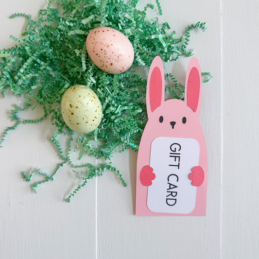 Gift card holder with bunny design next to Easter eggs and green grass on a white background
