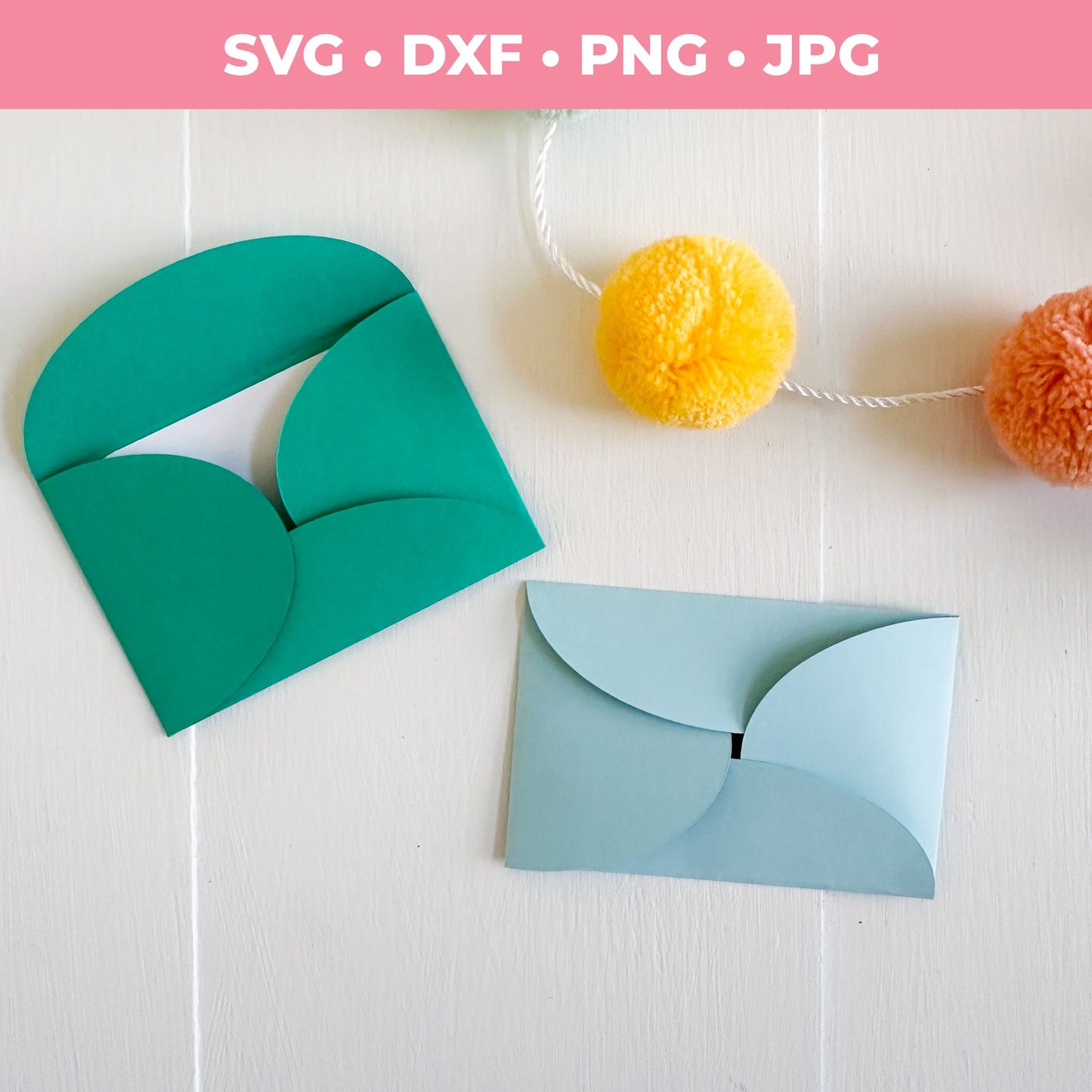 Two paper envelopes, one green and one blue, on a white surface with colorful pom-poms in the background.