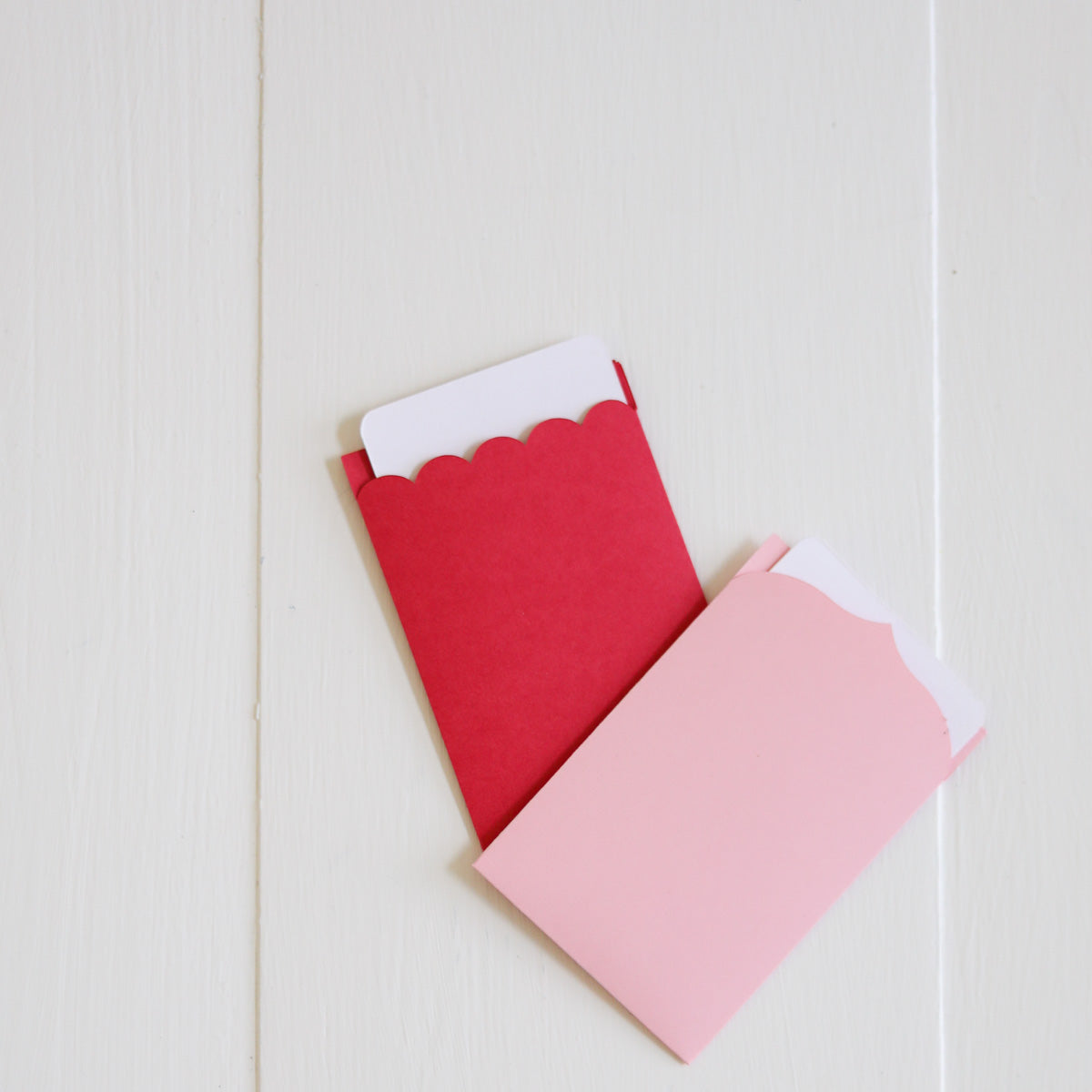 Two scalloped-edge envelopes, one red and one pink, on a light wooden surface.