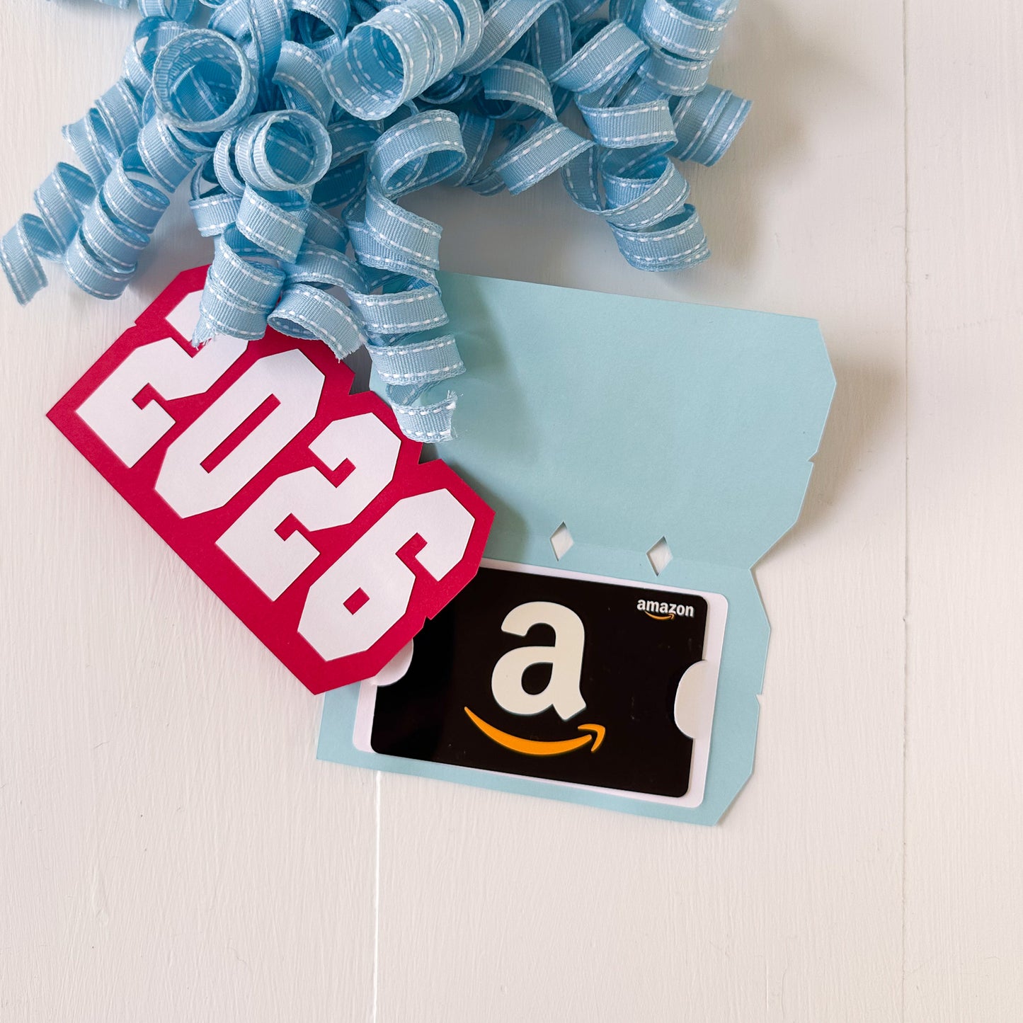 Amazon gift card with a blue ribbon and '2026' gift card holder on a light background