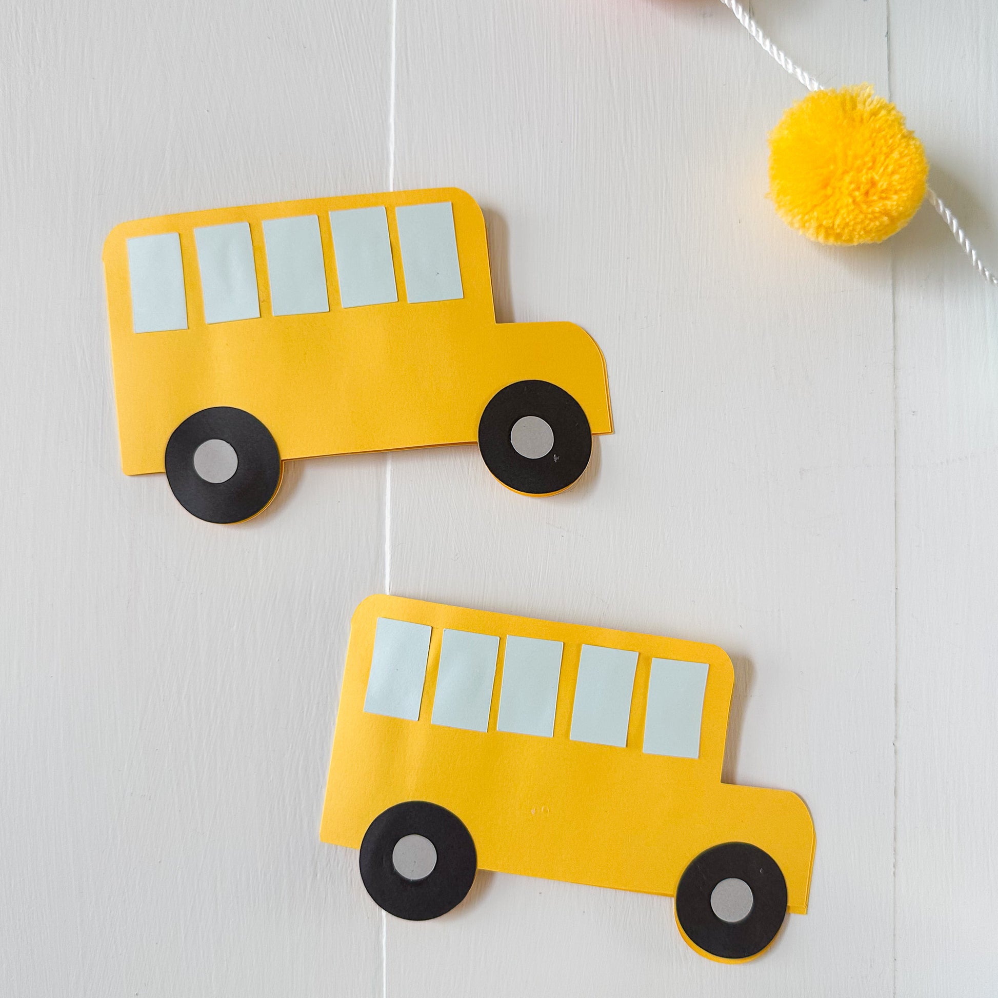 Two yellow paper school bus gift card holders on a white background with a yellow pom-pom garland.