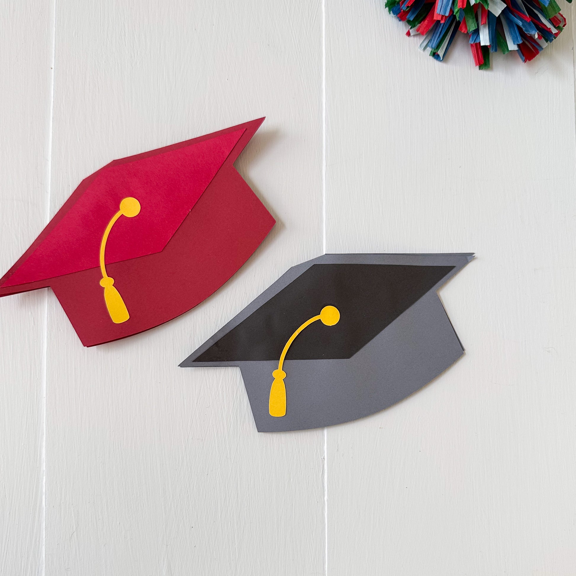 Red and gray graduation cap gift card holders with yellow tassels on a white wooden surface.
