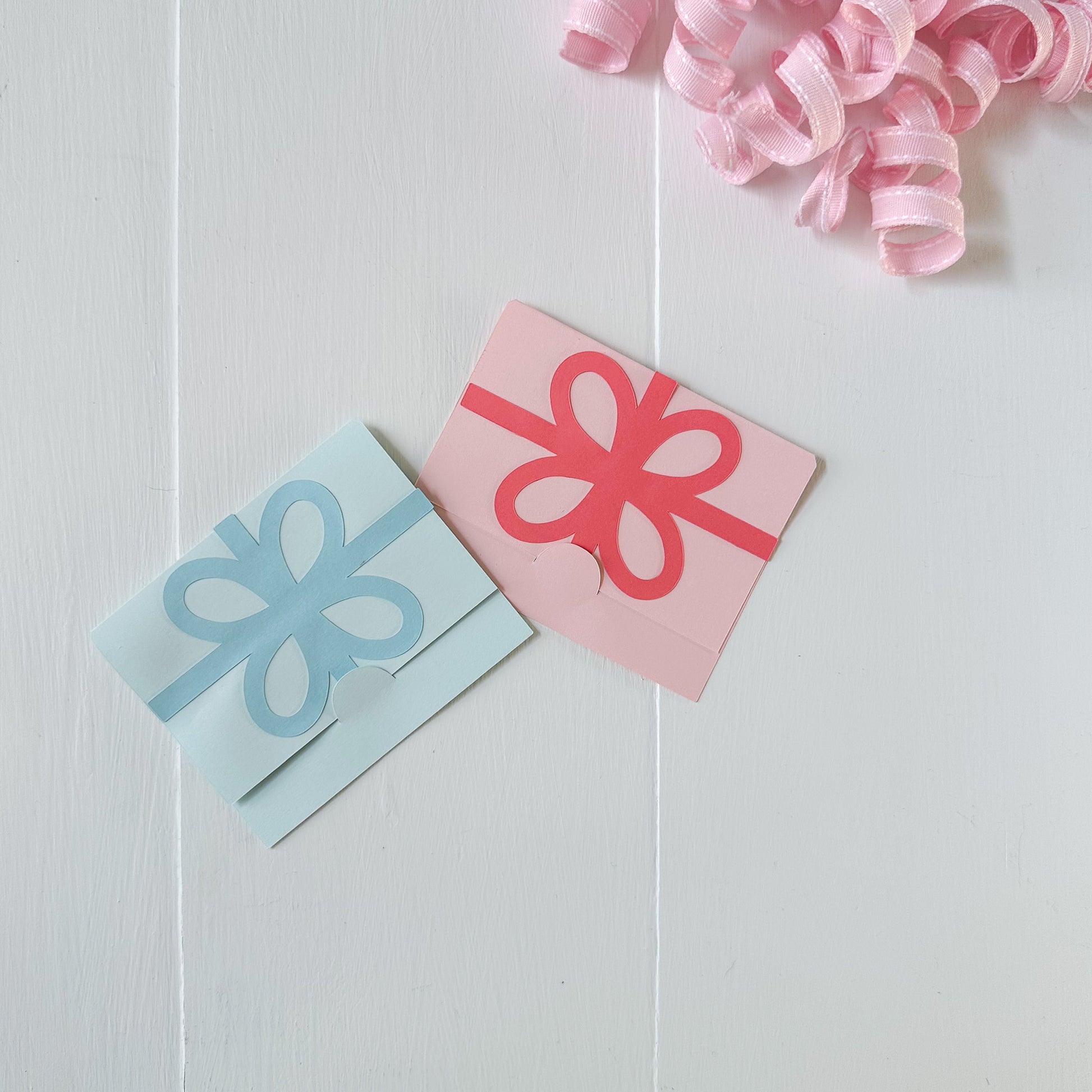 Two paper presents with bows, one blue and one pink, on a light wooden surface with pink ribbons.