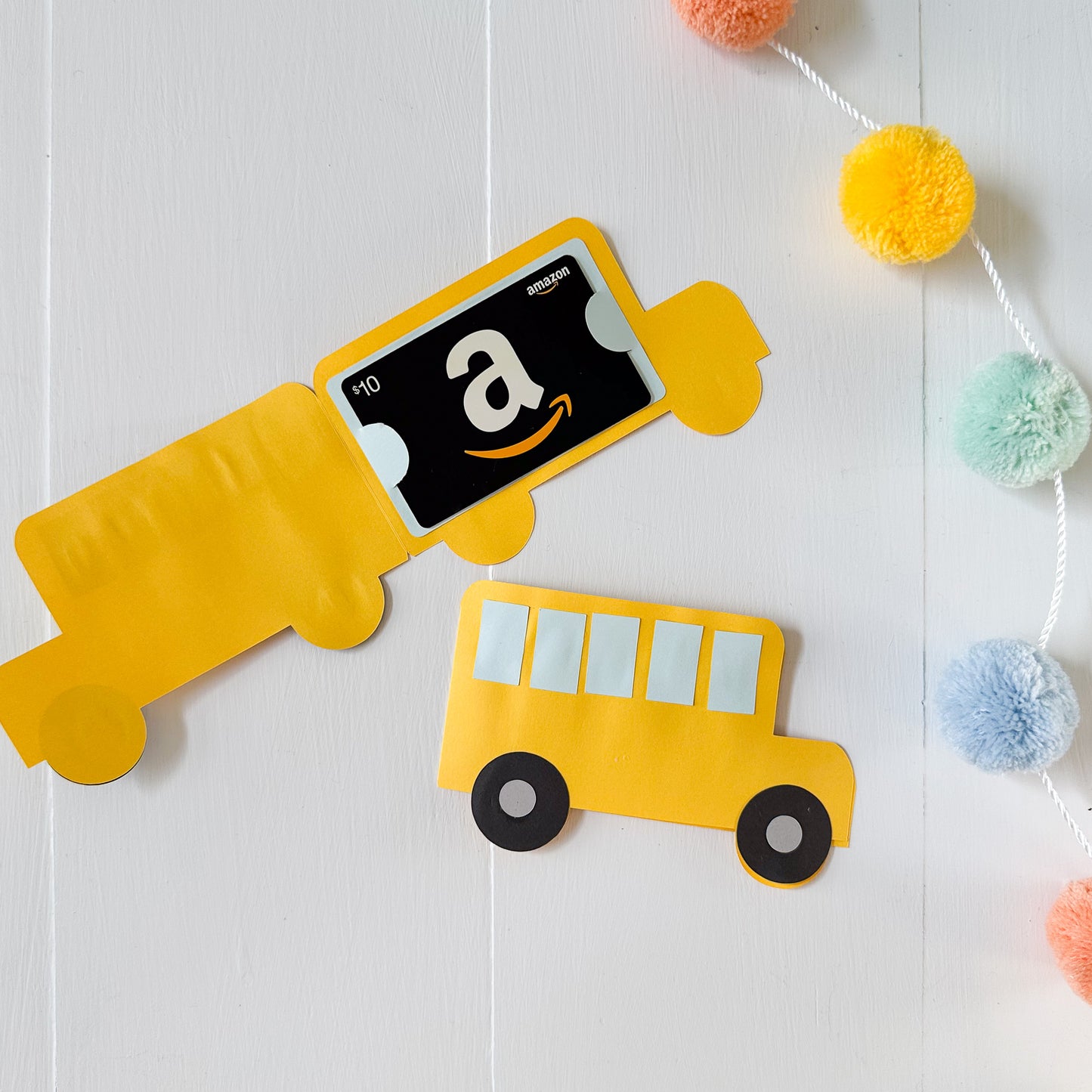 Yellow school bus-shaped Amazon gift card  holder on a white background with colorful pom-poms.