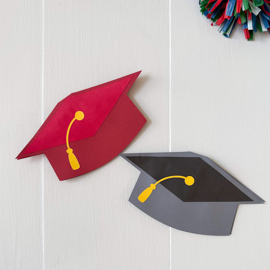 Red and black graduation cap gift card holders with yellow tassels on a light wooden surface